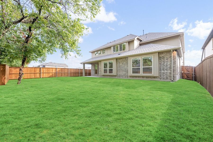 Backyard of two-story brick home with large green lawn, wood privacy fence, and covered patio.