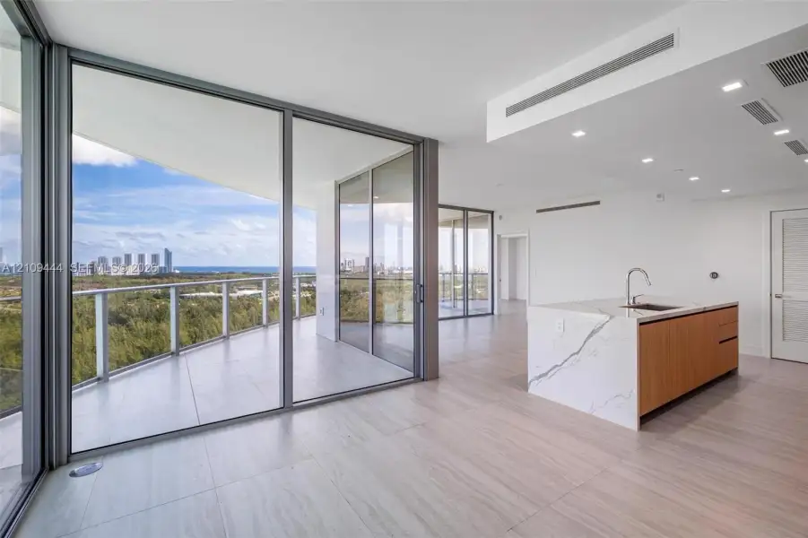 Floor-to-ceiling glass walls, expansive wraparound balcony, kitchen island with waterfall-edge stone countertop, wood-finish cabinetry, and neutral large-format tile flooring