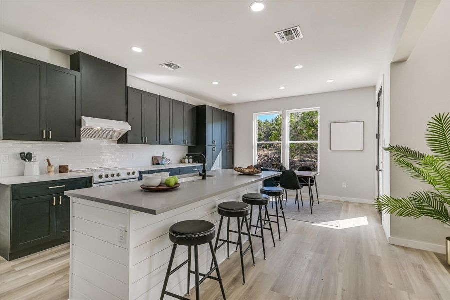 Kitchen with decorative backsplash, light wood-type flooring, a breakfast bar, a center island with sink, and under cabinet range hood