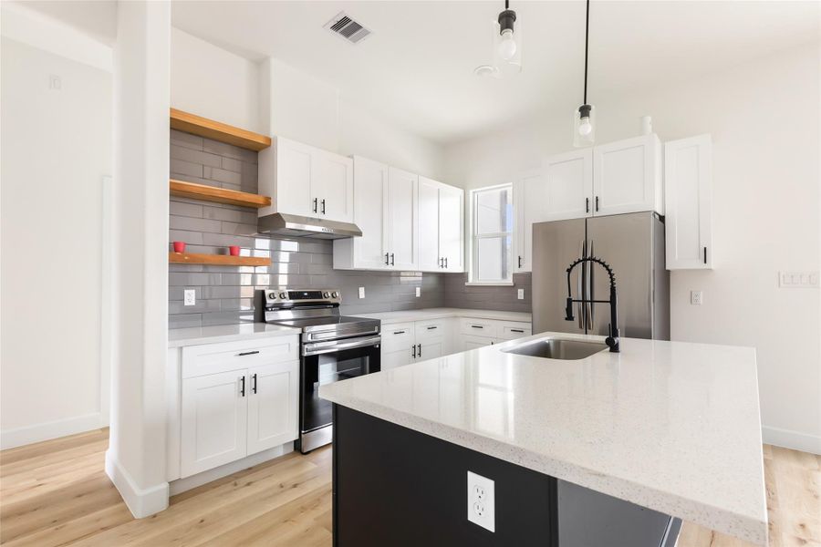 This modern kitchen features sleek white cabinetry, a gray subway tile backsplash, and stainless steel appliances. It includes a spacious island with a quartz countertop and a stylish pull-down faucet. Light wood flooring and open shelving add warmth and functionality.