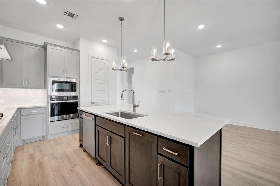 Kitchen featuring light wood-style floors, stainless steel appliances, an island with sink, suspended lighting, and backsplash Kitchen featuring light wood-style floors, stainless steel appliances, an island with sink, suspended lighting, and backsplash