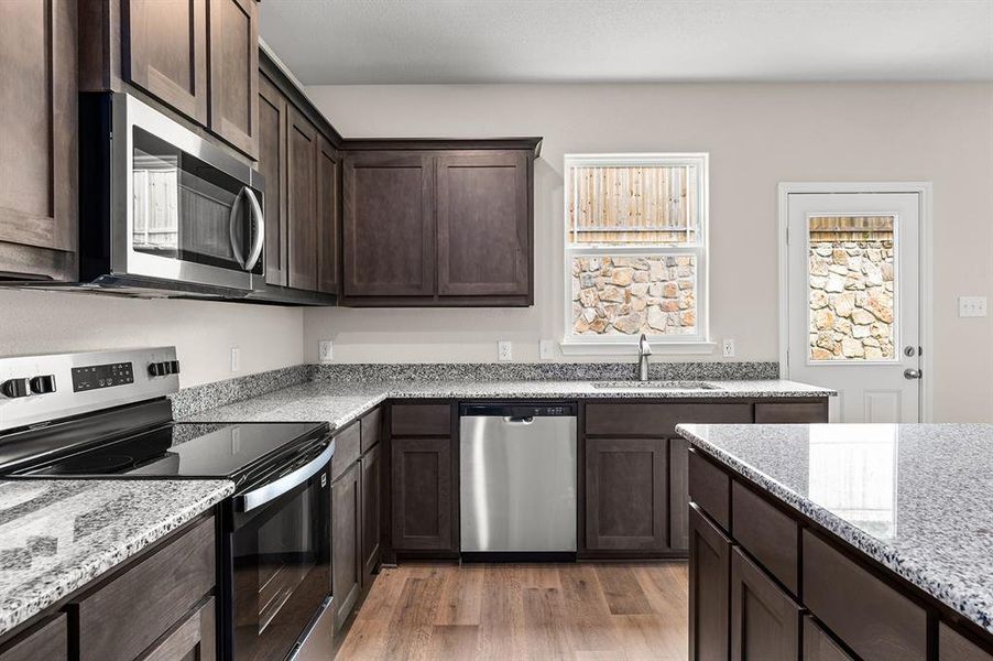 Kitchen with appliances with stainless steel finishes, light wood-style flooring, dark brown cabinetry, and light stone counters Kitchen with appliances with stainless steel finishes, light wood-style flooring, dark brown cabinetry, and light stone counters