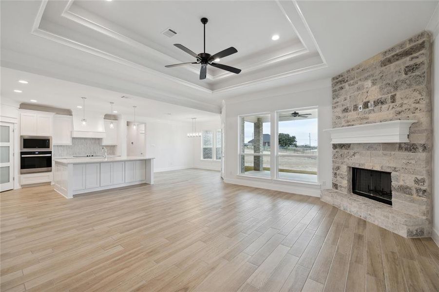 Unfurnished living room with a stone fireplace, ceiling fan with notable chandelier, a raised ceiling, and light hardwood / wood-style flooring