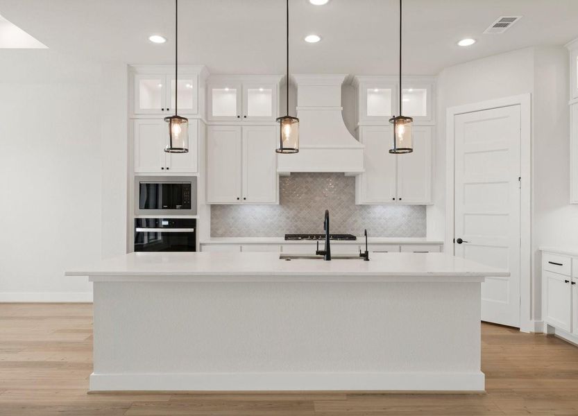 This photo showcases a beautiful kitchen with a clean,white design. It features a large island with a built-in sink, pendant lighting, stylish backsplash, quartz countertops & stainless appliances. The space is bright and open, with ample storage in the stacked cabinetry.