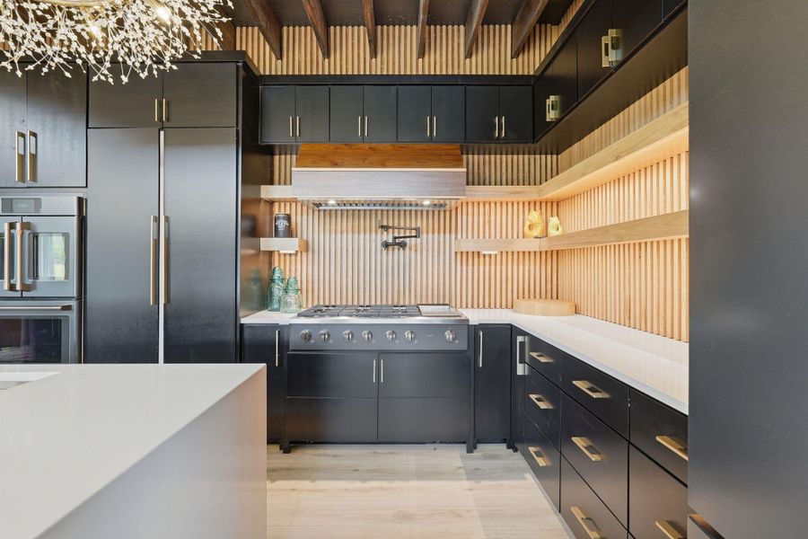 This modern kitchen features sleek black cabinetry with gold accents, a gas stove, and open shelving with wood accents. It includes a large refrigerator, built-in oven, and stylish lighting, creating a chic and functional space.