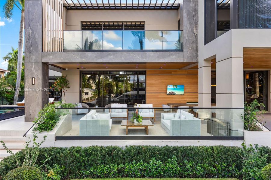 Elevated view of rear outdoor living terrace enclosed by glass railing, white woven lounge seating, wall-mounted TV on wood accent wall, stone façade upper level with glass balcony and pergola, pool at left, and manicured hedges throughout.