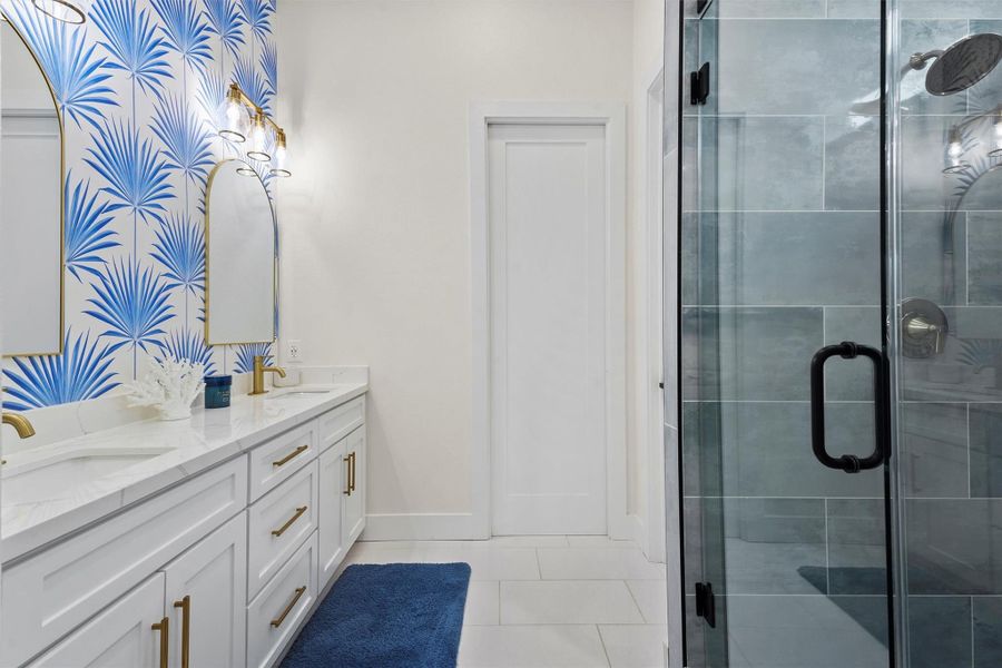 The primary bathroom features a modern design with a vibrant blue palm Serena & Lily wallpaper, white cabinetry, gold fixtures, and a sleek walk in glass shower. The room is bright and stylish, offering a fresh, contemporary feel.