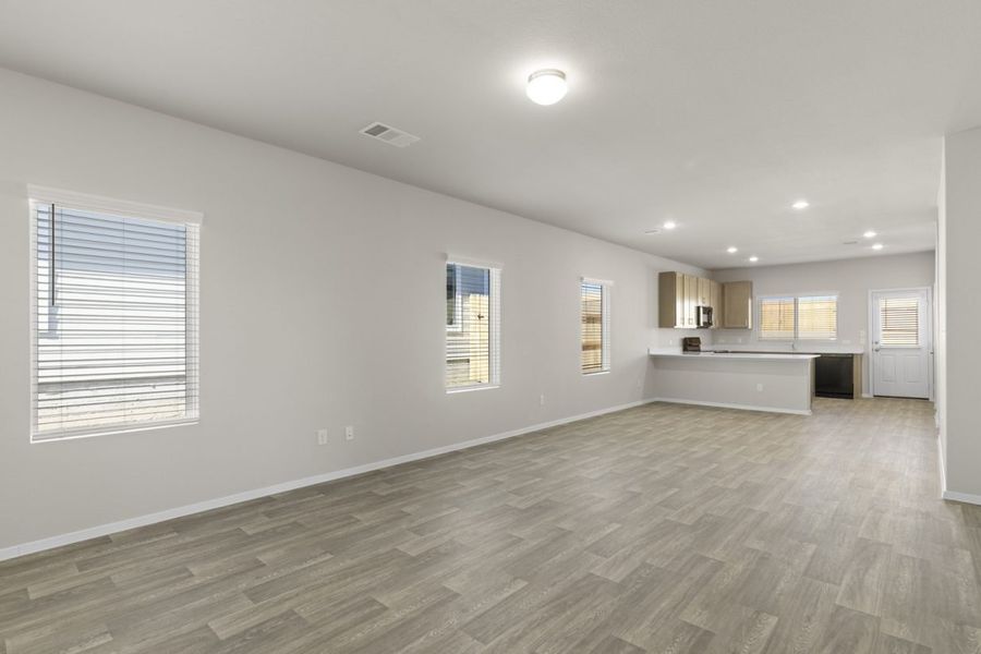 Image of a living room with beige walls, light brown wood-look flooring, windows with blinds, and an open kitchen in the distance Image of a living room with beige walls, light brown wood-look flooring, windows with blinds, and an open kitchen in the distance