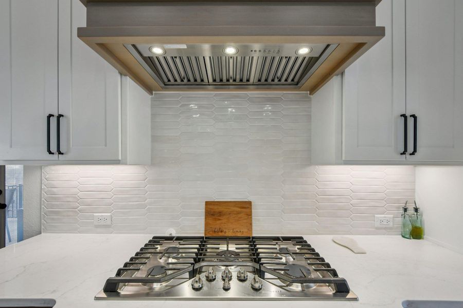 The kitchen features a stainless steel gas cooktop, a stylish range hood with wood and metal accents, white cabinetry with black hardware, and a white tile backsplash The kitchen features a stainless steel gas cooktop, a stylish range hood with wood and metal accents, white cabinetry with black hardware, and a white tile backsplash