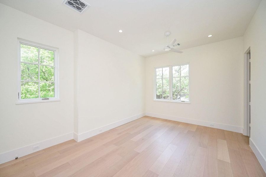 Bedroom two on second floor, bright, spacious room features light wood floors, neutral walls, and large windows that offer abundant natural light and views of greenery. It's equipped with recessed lighting and a ceiling fan for comfort.