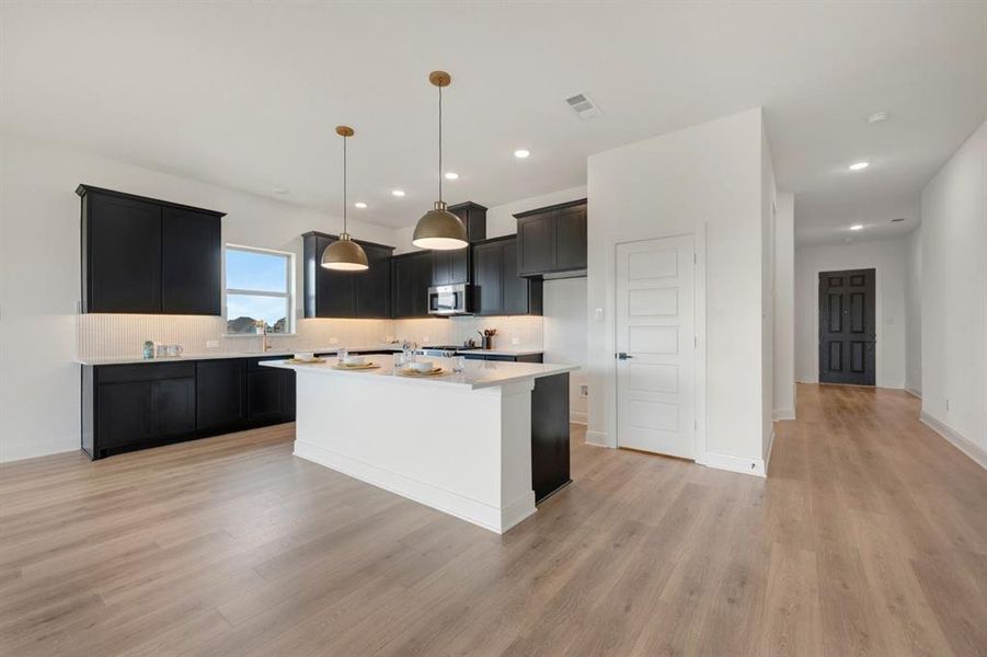 Kitchen featuring tasteful backsplash, a kitchen island, pendant lighting, stainless steel microwave, and light wood-style flooring Kitchen featuring tasteful backsplash, a kitchen island, pendant lighting, stainless steel microwave, and light wood-style flooring