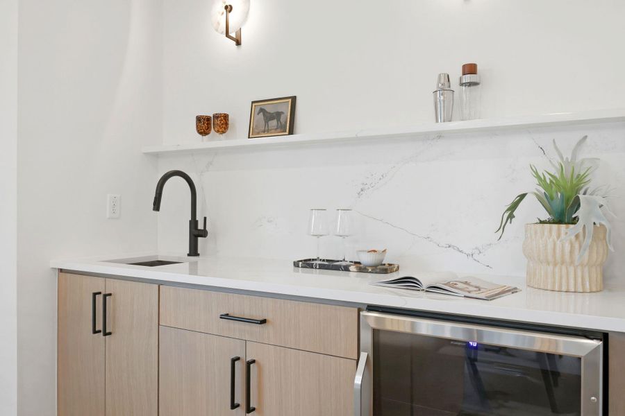 Wet bar featuring light wood-finish cabinetry, a white countertop with a matching backsplash, a stainless steel beverage refrigerator, a black gooseneck faucet, and an undermount sink