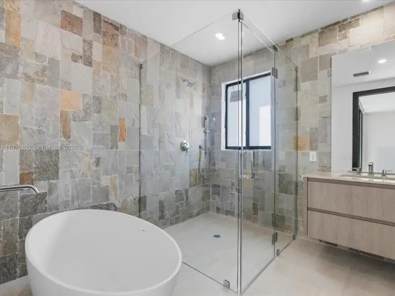 Freestanding soaking tub, frameless glass-enclosed shower, natural stone-tiled walls, wall-mounted floating vanity, and large-format tile flooring Freestanding soaking tub, frameless glass-enclosed shower, natural stone-tiled walls, wall-mounted floating vanity, and large-format tile flooring