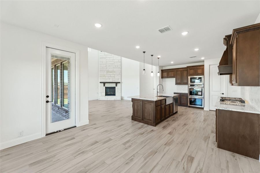 Open kitchen with dark wood cabinets, center island, and stone fireplace visible in adjoining living area with light wood floors Open kitchen with dark wood cabinets, center island, and stone fireplace visible in adjoining living area with light wood floors