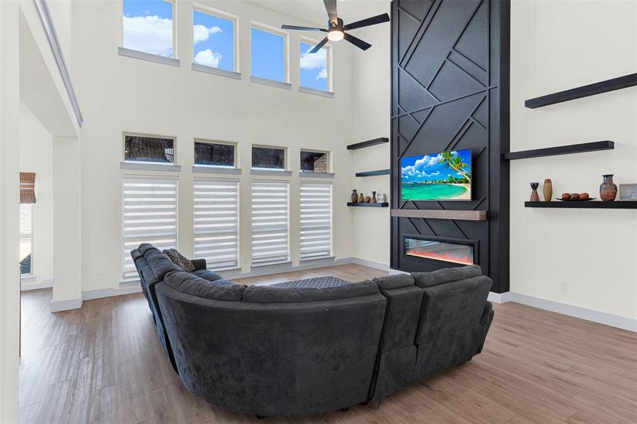 Living room featuring a large fireplace, a ceiling fan, a high ceiling, and light wood-style flooring Living room featuring a large fireplace, a ceiling fan, a high ceiling, and light wood-style flooring