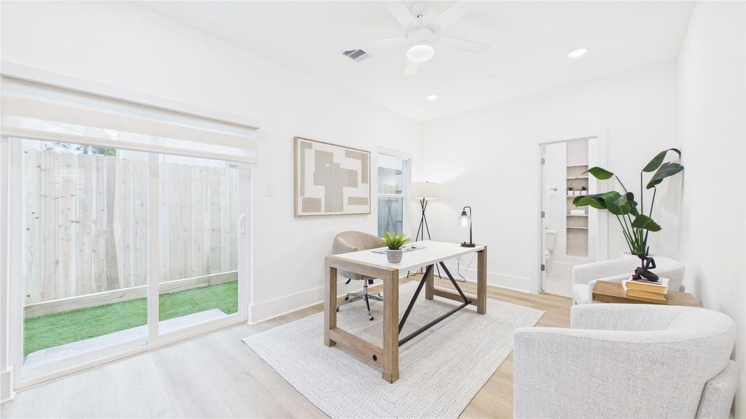 Bright, modern home office with sliding glass doors opening to a small fenced yard. Features include light wood flooring, a ceiling fan, and a minimalist decor with a desk, chairs, and greenery. Perfect for a cozy, productive workspace.