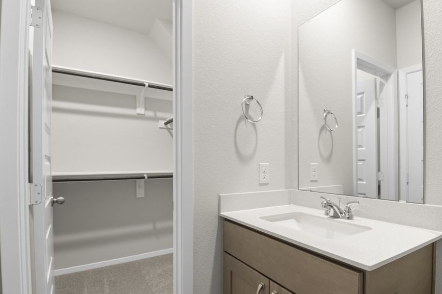 Image of a primary bathroom with brown cabinets and a white vanity with mirror and a connected walk-in closet