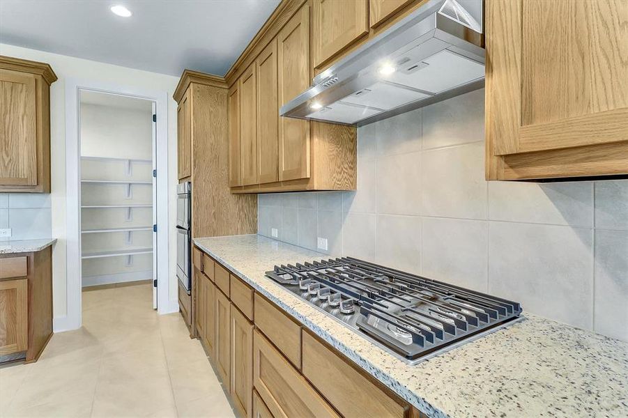Kitchen featuring natural wood cabinetry, light speckled countertops, a built-in gas range with an overhead stainless steel vent hood, and an expansive walk-in pantry Kitchen featuring natural wood cabinetry, light speckled countertops, a built-in gas range with an overhead stainless steel vent hood, and an expansive walk-in pantry