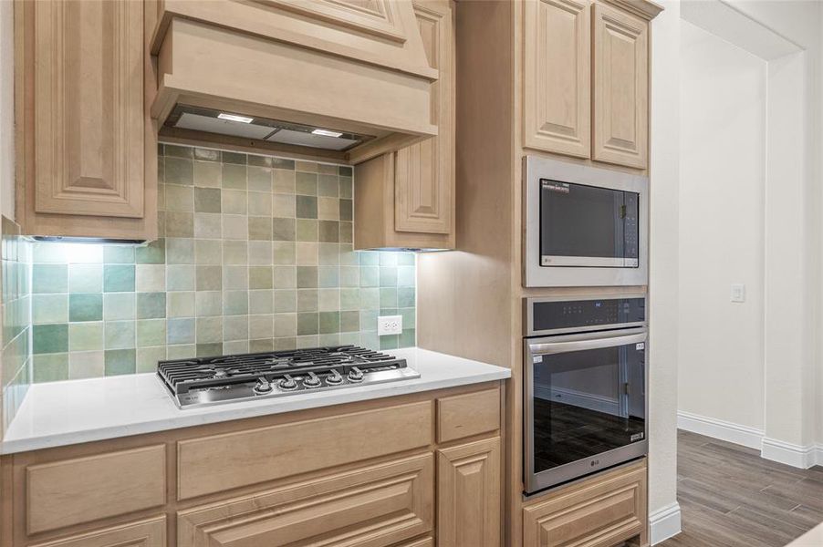 Kitchen with custom range hood, appliances with stainless steel finishes, decorative backsplash, wood finished floors, and light brown cabinetry Kitchen with custom range hood, appliances with stainless steel finishes, decorative backsplash, wood finished floors, and light brown cabinetry