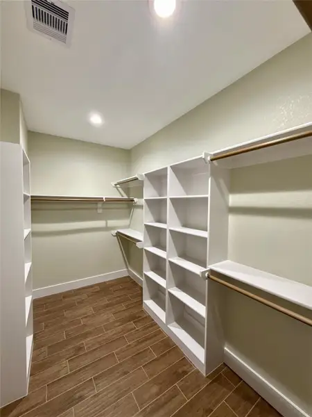 This spacious walk-in closet features ample shelving and hanging space, a light neutral color scheme, and wood-look tile flooring for a warm, organized, and inviting storage solution.