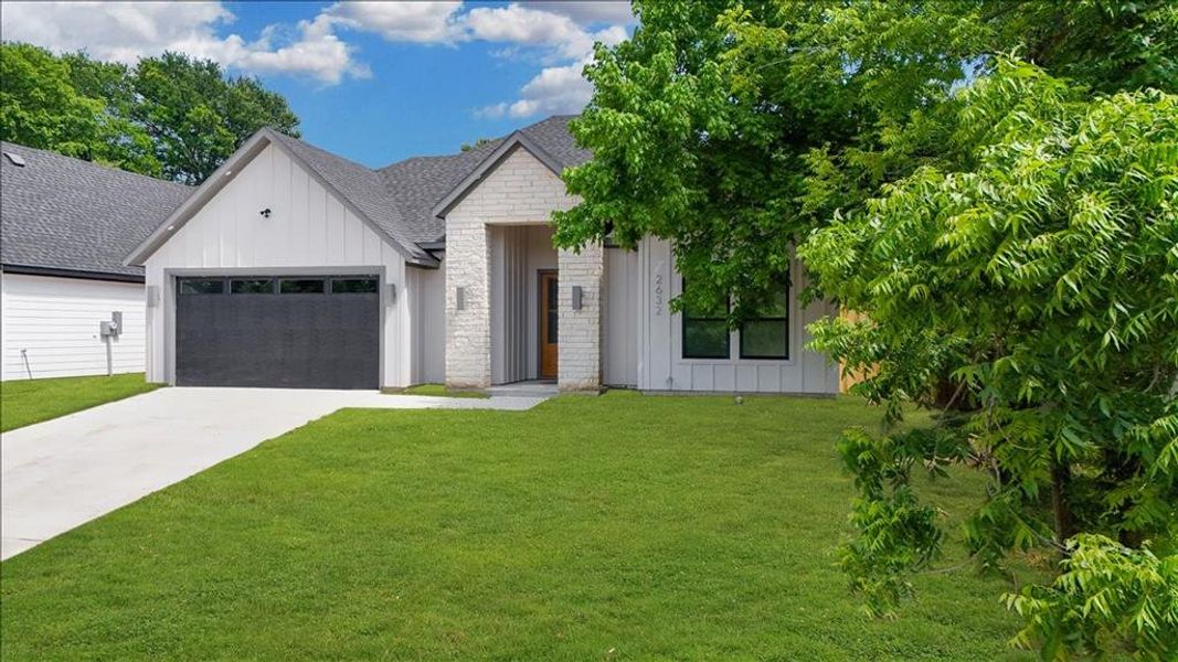 Modern farmhouse-style exterior featuring board and batten siding, light-colored brick accents, a dark garage door with window inserts, a concrete driveway, and a front lawn with mature trees Modern farmhouse-style exterior featuring board and batten siding, light-colored brick accents, a dark garage door with window inserts, a concrete driveway, and a front lawn with mature trees