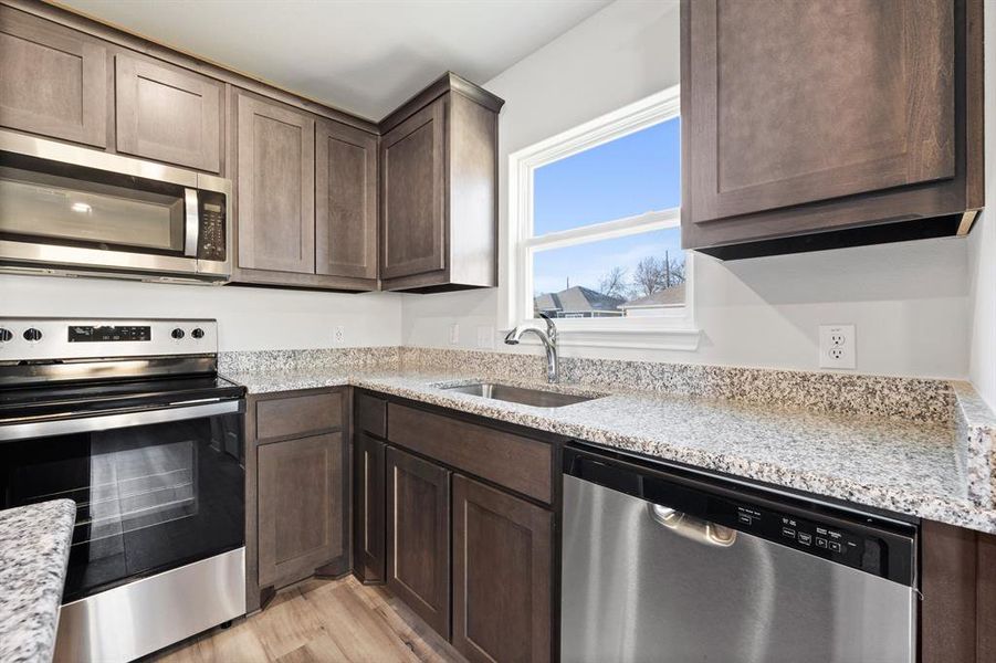 Kitchen with stainless steel appliances, light hardwood / wood-style floors, dark brown cabinets, sink, and light stone counters