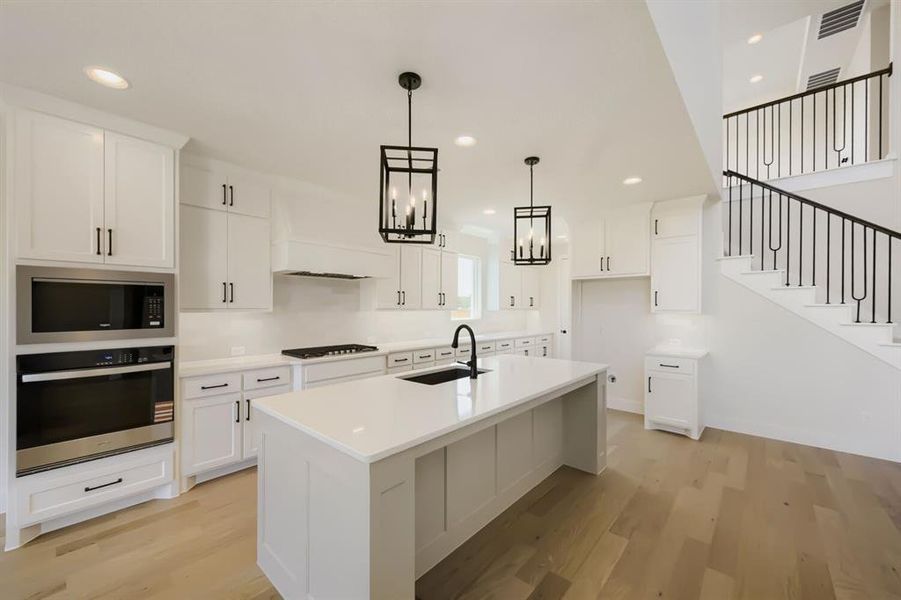 Gourmet kitchen featuring white shaker cabinetry, an expansive center island with an undermount sink and matte black faucet, stainless steel built-in appliances, and light wood-finish flooring