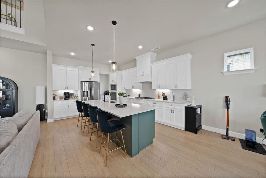 Modern kitchen featuring an expansive central island with seating, white shaker cabinetry, stainless steel appliances, and light wood-finish flooring Modern kitchen featuring an expansive central island with seating, white shaker cabinetry, stainless steel appliances, and light wood-finish flooring