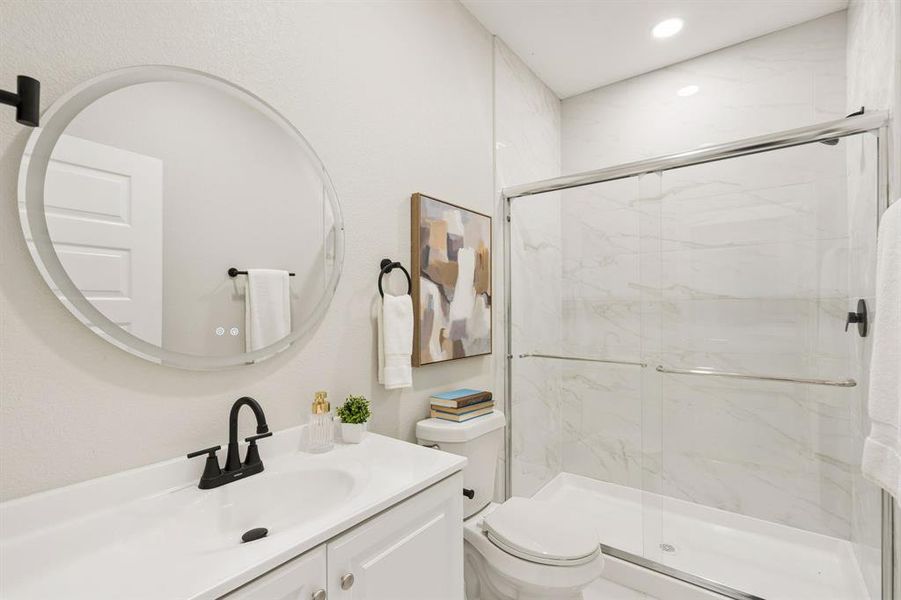 Bathroom featuring a glass-enclosed shower with white tiled walls, a white vanity with integrated sink, a circular illuminated mirror, and matte black fixtures Bathroom featuring a glass-enclosed shower with white tiled walls, a white vanity with integrated sink, a circular illuminated mirror, and matte black fixtures