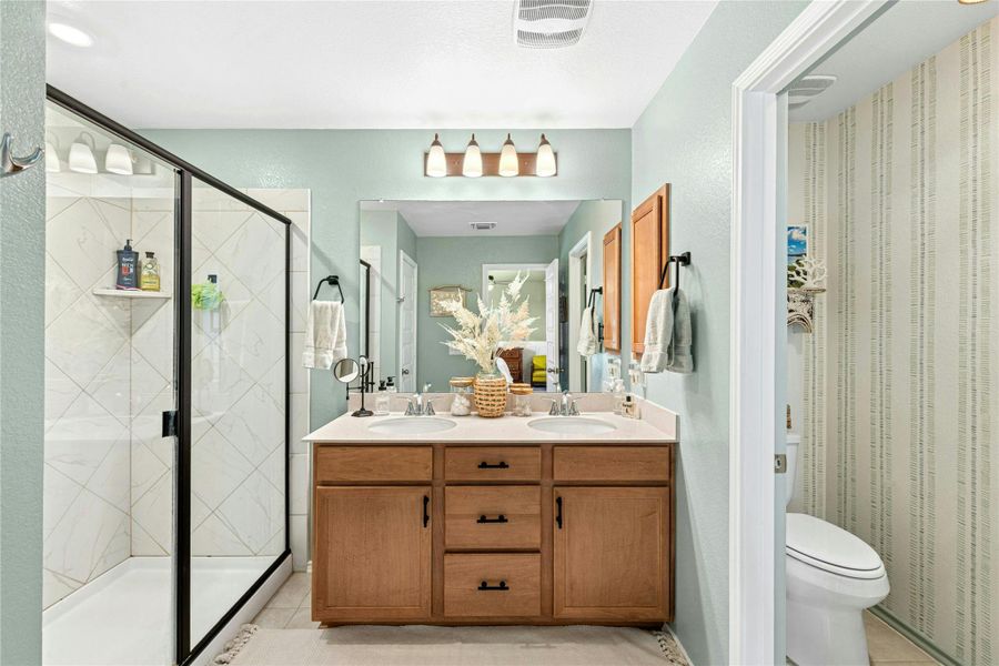 The primary bathroom offers a refined retreat, showcasing a double vanity, light tile-patterned flooring, an extra-large tiled shower with sleek glass doors, and tasteful decorative accents throughout.
