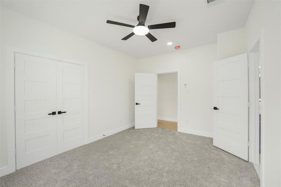 This room features plush carpeting and neutral walls, creating a cozy atmosphere. It has a modern ceiling fan and ample natural light. The space includes a double-door closet and an additional door leading to another area, offering convenience and functionality.