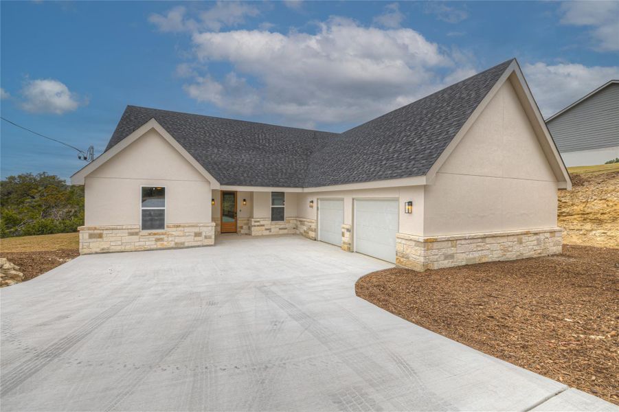 View of front of property with stucco siding, stone siding, an attached garage, a shingled roof, and concrete driveway