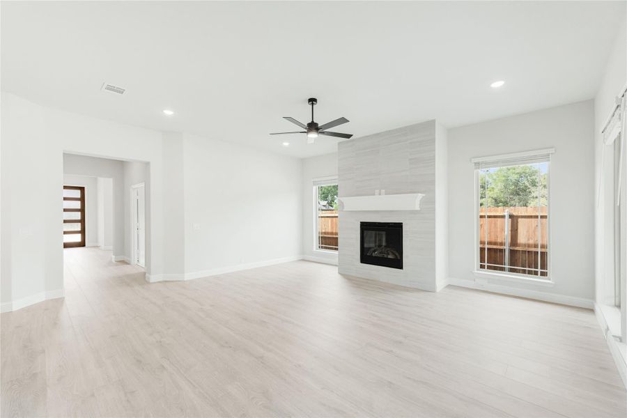 Open living room with light wood floors, tiled fireplace, ceiling fan, and large windows