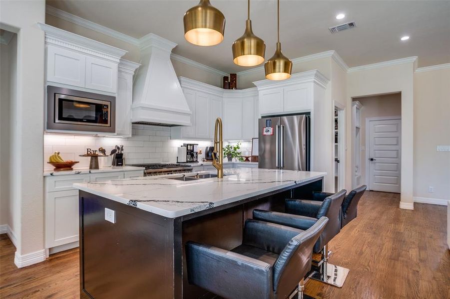 Kitchen featuring premium range hood, white cabinets, a center island with sink, decorative light fixtures, and stainless steel appliances