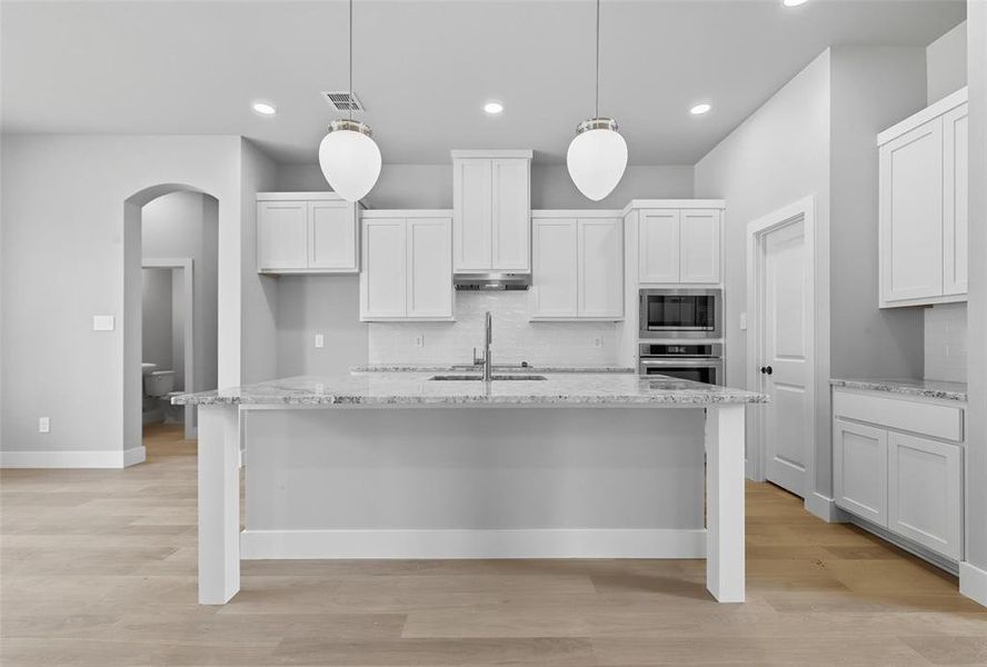 Kitchen featuring arched walkways, light stone countertops, an island with sink, light wood-style floors, and decorative light fixtures