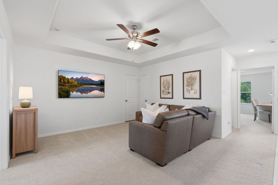 This spacious, carpeted living room features a ceiling fan and neutral walls, providing a cozy atmosphere. It includes a comfortable brown leather sofa, wall art, and a flat-screen TV. A doorway leads to a bright adjacent room, ideal for a home office or dining area.