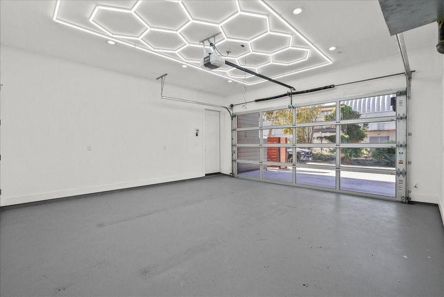 This photo shows a modern, spacious garage with sleek hexagonal ceiling lights and a large, glass-panel garage door letting in natural light. Ideal for parking or a workshop space. This photo shows a modern, spacious garage with sleek hexagonal ceiling lights and a large, glass-panel garage door letting in natural light. Ideal for parking or a workshop space.