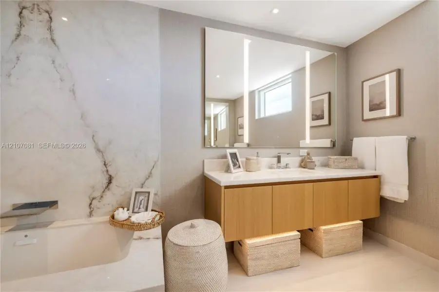 Floating wood-finish vanity with a stone countertop, wall-mounted faucet, integrated mirror with vertical LED lighting, and full-height stone tile surround at the soaking tub Floating wood-finish vanity with a stone countertop, wall-mounted faucet, integrated mirror with vertical LED lighting, and full-height stone tile surround at the soaking tub