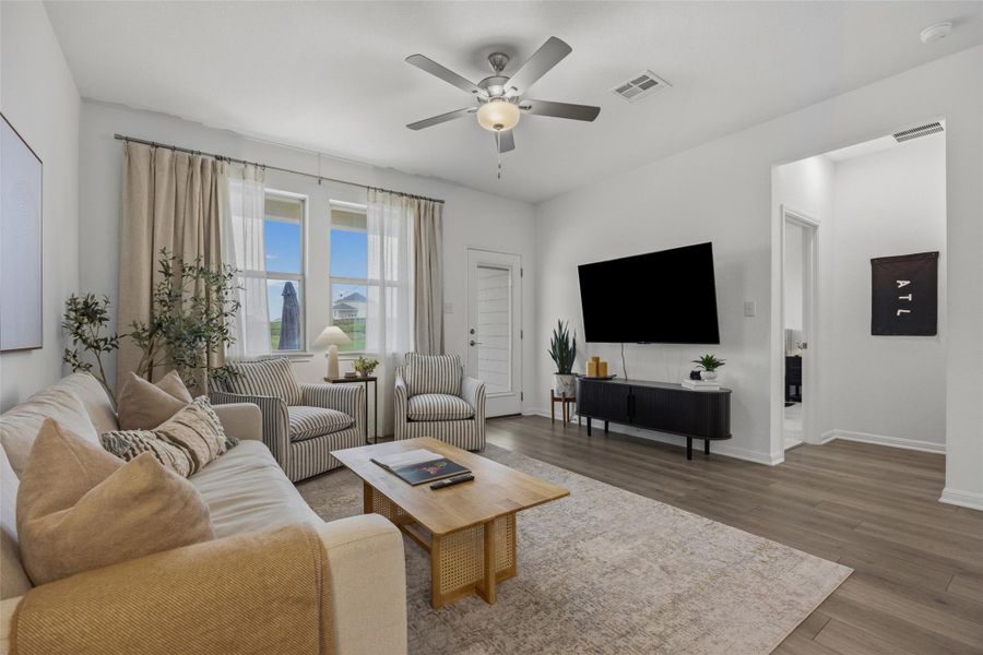 Bright living area with wood-finish flooring, a ceiling fan with integrated lighting, and multiple windows providing natural illumination