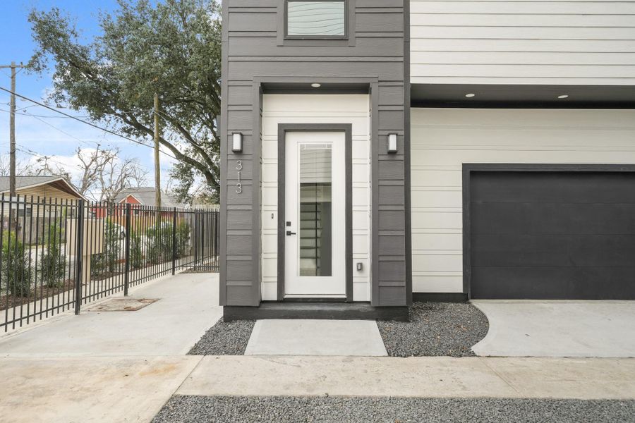 The front of this home features a cement board facade accented by recessed lights under the garage entry, while two coach lights frame the 8-foot glass front door with integrated blinds. The wrought iron gate provides access to a backyard finished with black gravel. The front of this home features a cement board facade accented by recessed lights under the garage entry, while two coach lights frame the 8-foot glass front door with integrated blinds. The wrought iron gate provides access to a backyard finished with black gravel.