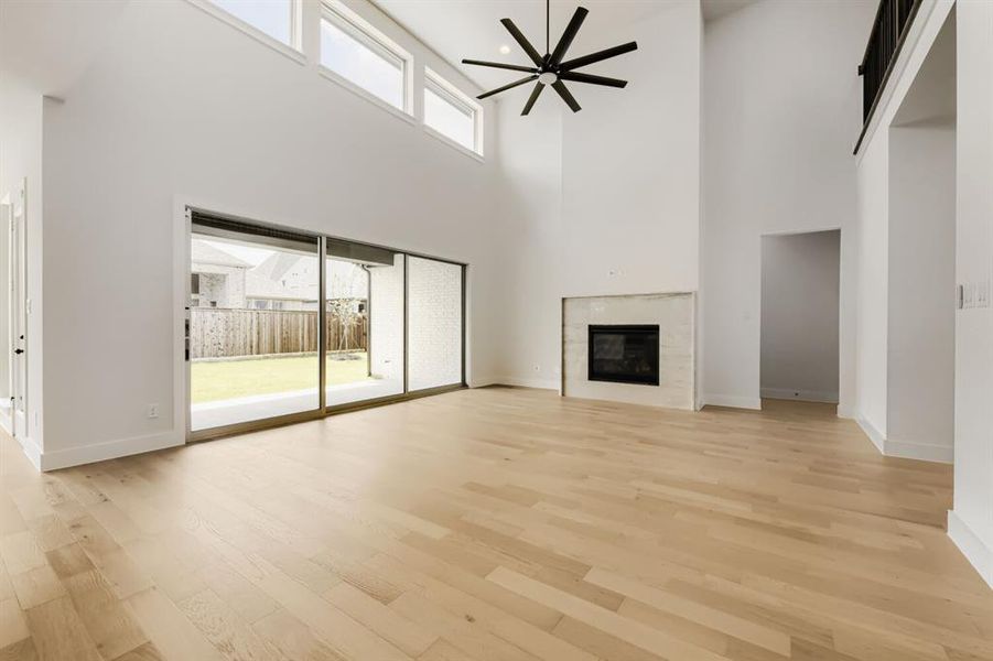 Double-height interior featuring light wood-finish flooring, a contemporary fireplace with a light-toned surround, a large black ceiling fan, and extensive natural light from transom windows and a multi-panel sliding glass door opening to a private backyard with a patio Double-height interior featuring light wood-finish flooring, a contemporary fireplace with a light-toned surround, a large black ceiling fan, and extensive natural light from transom windows and a multi-panel sliding glass door opening to a private backyard with a patio
