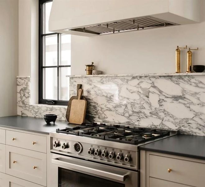 Gourmet kitchen featuring a stainless steel range with six burners, a matching built-in range hood, full-height marble backsplash, and light-toned cabinetry with brass hardware