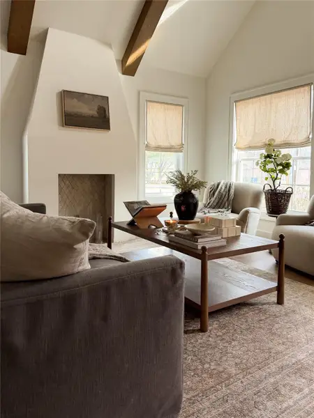 Vaulted ceiling with exposed wood beams, a fireplace with herringbone pattern firebox, a textured surround, and two large windows featuring natural fiber faux Roman shades Vaulted ceiling with exposed wood beams, a fireplace with herringbone pattern firebox, a textured surround, and two large windows featuring natural fiber faux Roman shades