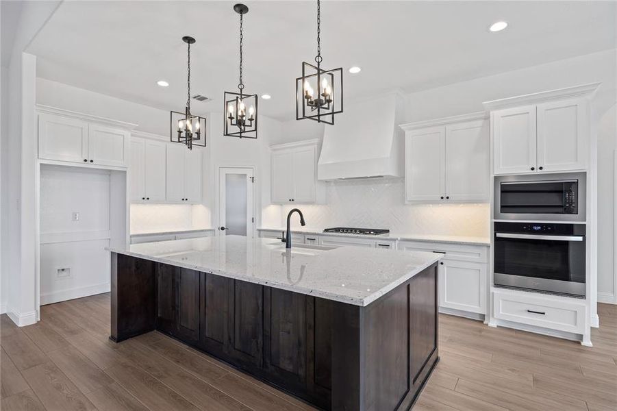 Kitchen with dual tone cabinets, stainless steel appliances, a center island with sink, suspended lighting, and light wood-type flooring