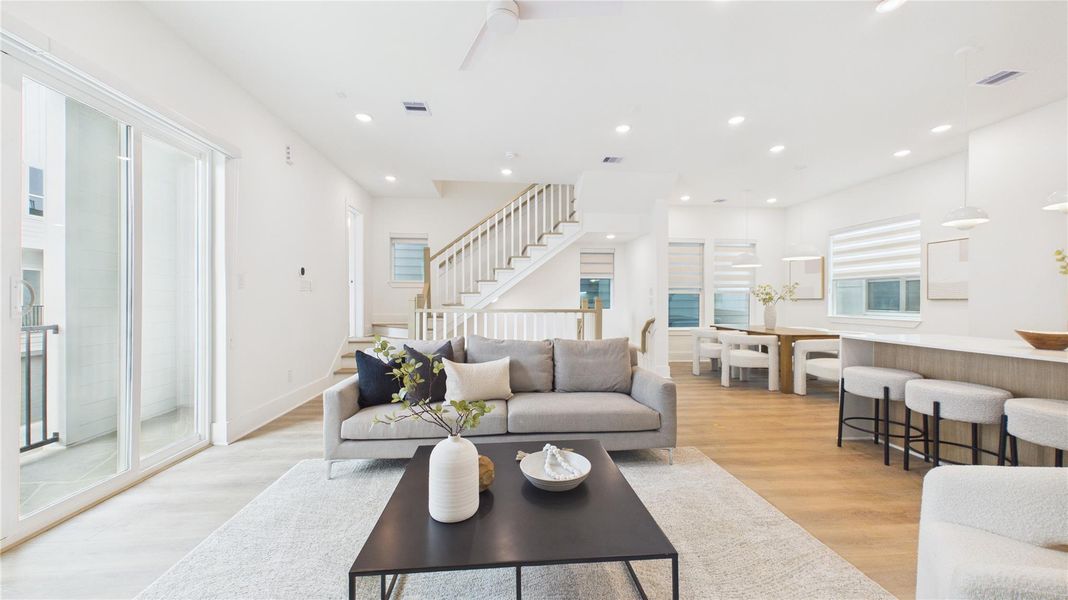 This photo showcases a bright, open-plan living area with modern decor. It features large sliding glass doors, a cozy sitting area, and a sleek kitchen with a breakfast bar. The space is enhanced by wood flooring, recessed lighting, and a contemporary staircase leading to the upper level, creating a welcoming and stylish ambiance.