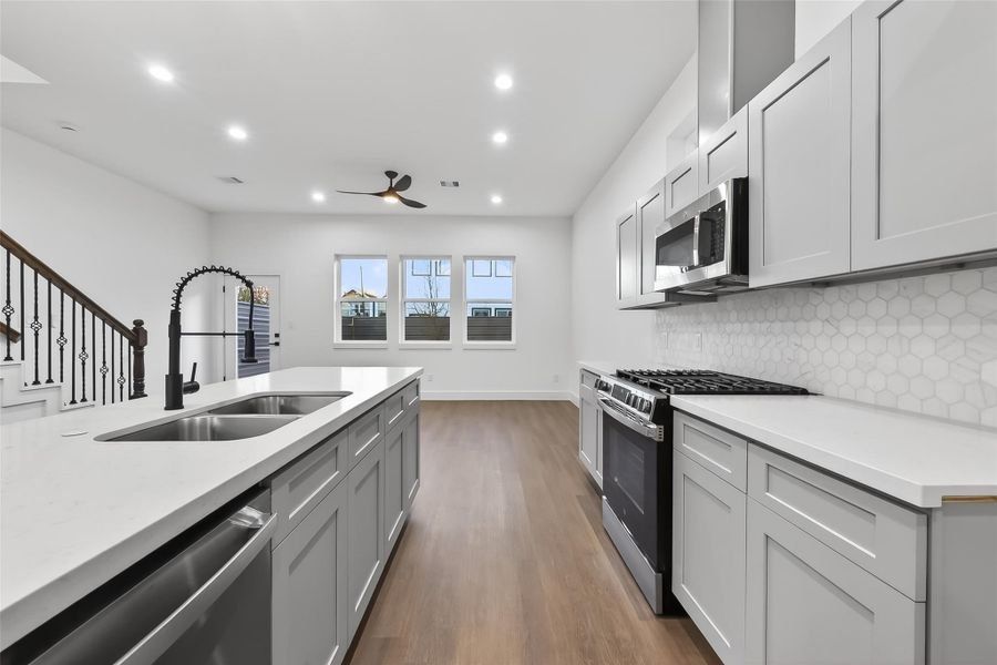 This modern kitchen features sleek gray cabinetry, a spacious island with dual sinks, and stainless steel appliances. The open layout is enhanced by wood flooring, a stylish backsplash, and ample natural light from the large windows.