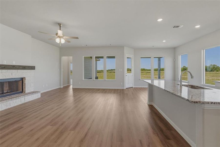 Open-concept living area featuring wood-finish flooring, a white brick fireplace with a wood mantle, and an integrated kitchen island with a granite-style countertop and undermount sink Open-concept living area featuring wood-finish flooring, a white brick fireplace with a wood mantle, and an integrated kitchen island with a granite-style countertop and undermount sink