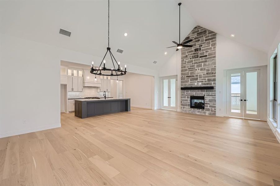 Unfurnished living room with a stone fireplace, high vaulted ceiling, ceiling fan with notable chandelier, light hardwood / wood-style floors, and french doors