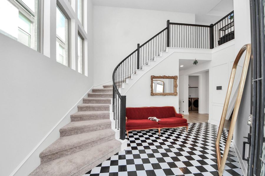 This photo showcases a bright, modern entryway featuring a striking black-and-white checkered floor, a grand rotunda staircase with black railings, and large windows for natural light. A red sofa adds a pop of color, complemented by a stylish gold-framed mirror. This photo showcases a bright, modern entryway featuring a striking black-and-white checkered floor, a grand rotunda staircase with black railings, and large windows for natural light. A red sofa adds a pop of color, complemented by a stylish gold-framed mirror.