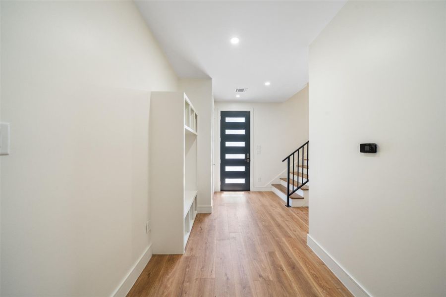 Well-lit entryway with engineered white oak wood flooring, a sleek black front door with horizontal glass panels, and a staircase with a black railing. The walls are painted in a light color, creating a bright and inviting atmosphere.